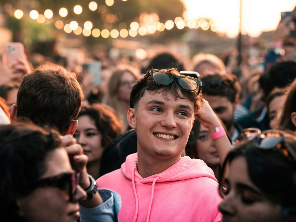 Young man in pink sweatshirt smiles in a crowd.