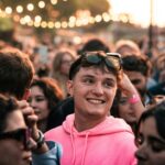 Young man in pink sweatshirt smiles in a crowd.