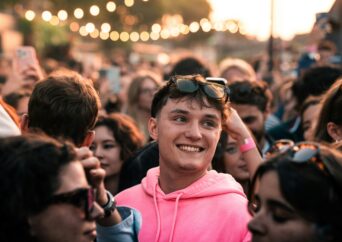 Young man in pink sweatshirt smiles in a crowd.