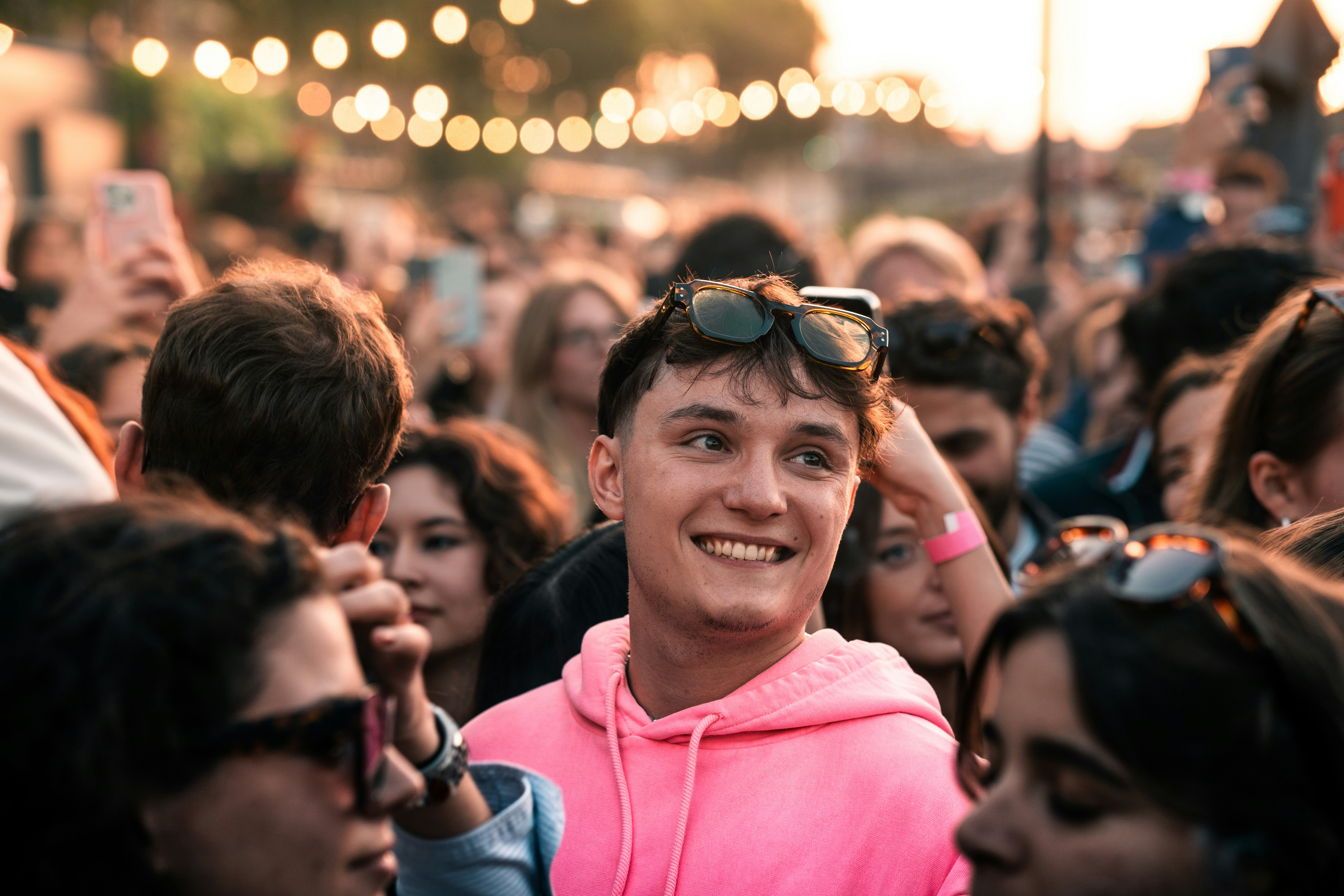 Young man in pink sweatshirt smiles in a crowd.